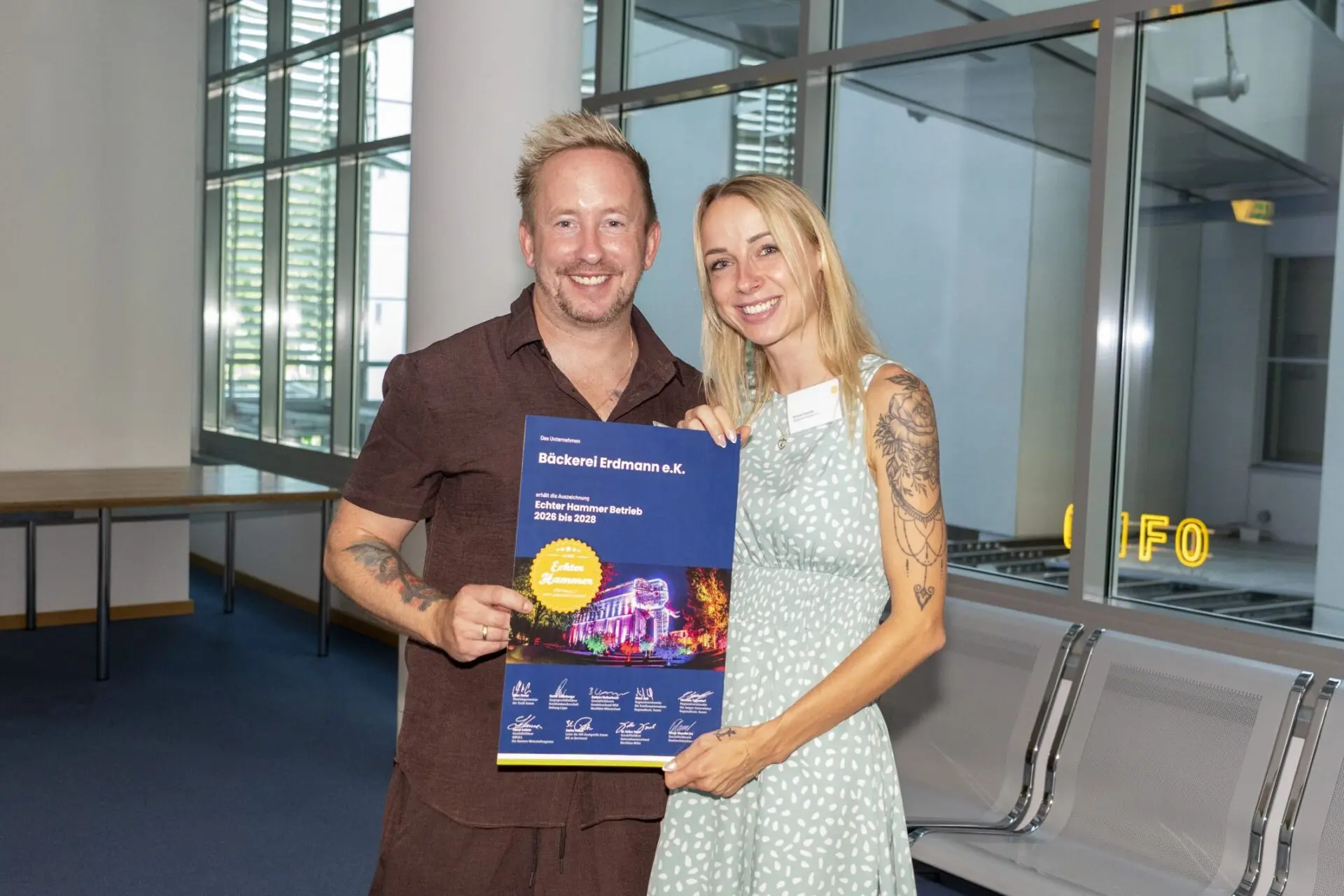 Ein Mann und eine Frau l&auml;cheln und halten ein blaues Zertifikat f&uuml;r B&auml;ckerei Erdmann e.K. mit der Auszeichnung &bdquo;Echter Hammer Betrieb 2026 bis 2028&ldquo;.